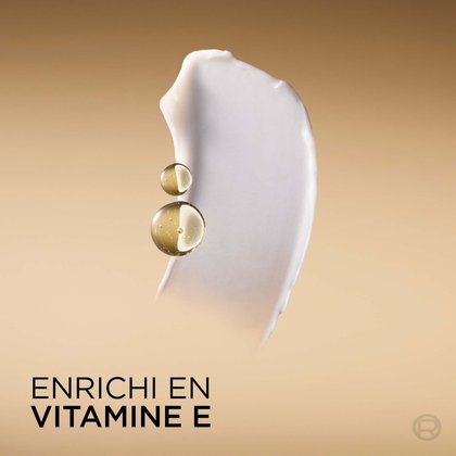 Un gros plan d'une trace de revitalisant blanc crémeux et lisse. Deux gouttelettes de vitamine E liquide dorée sont superposées sur la crème. Le texte en bas indique : « INFUSÉ DE VITAMINE E ».