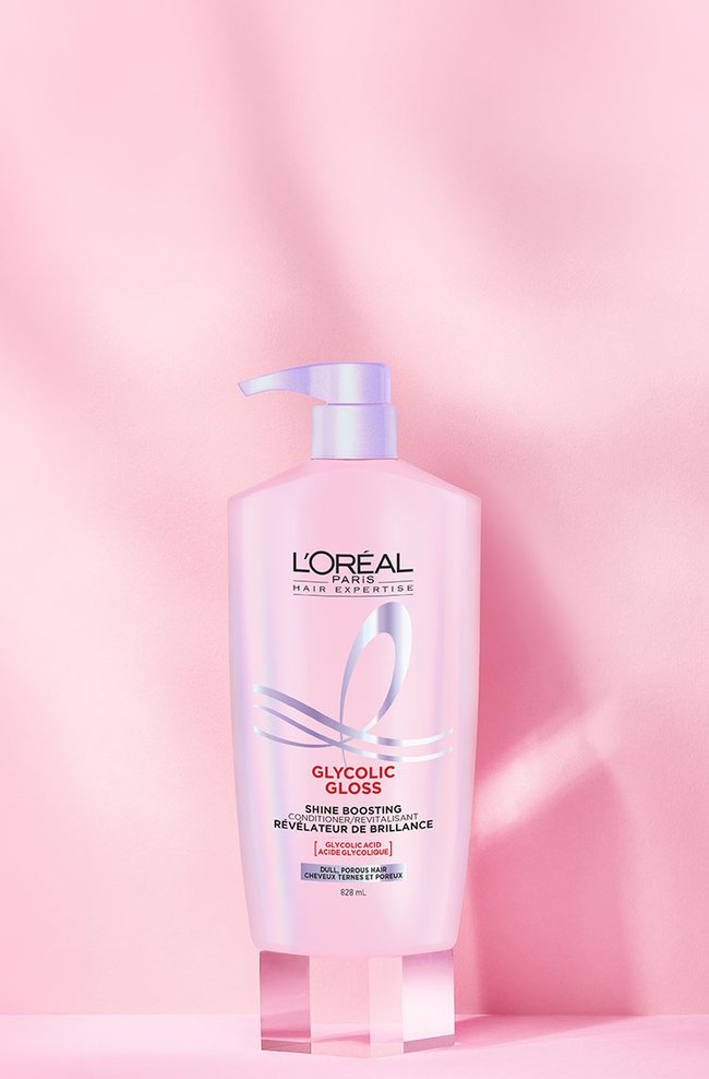 A L'Oréal Glycolic Gloss shampoo/conditioner pump bottle resting on a clear block, on a pink background with shadows.