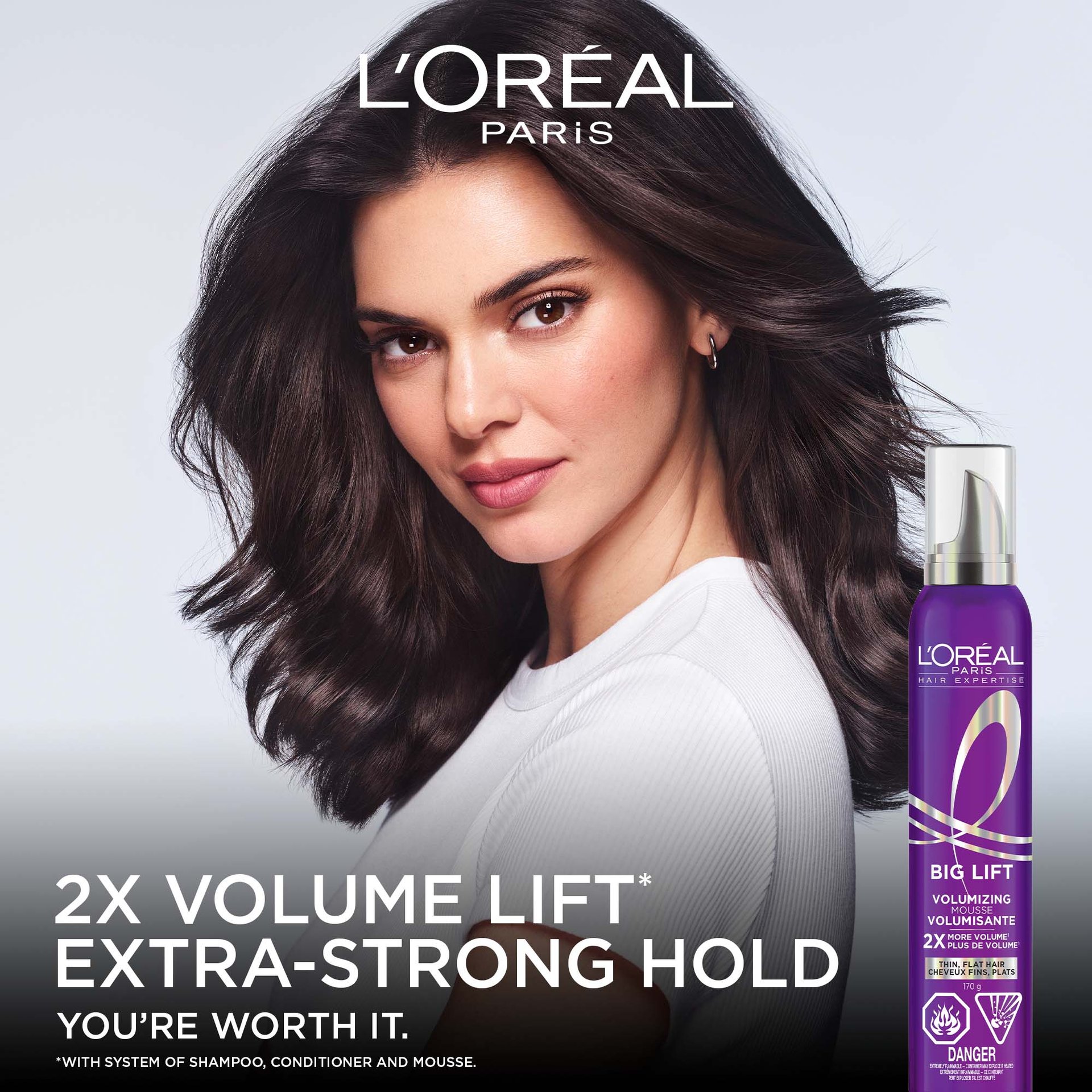 A female model with voluminous, wavy dark brown hair posing on a dark background next to a purple can of Hair Expertise Collagen Peptide Lifter Big Lift mousse, with text: "2X VOLUME LIFT, EXTRA-STRONG HOLD".