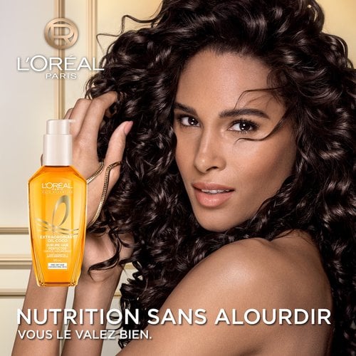 Une femme aux cheveux bouclés foncés, volumineux et brillants sourit à côté d'un flacon de L'Oréal Paris Huile Extraordinaire. En bas, le texte indique « Nutrition en toute légèreté pour cheveux fins ».