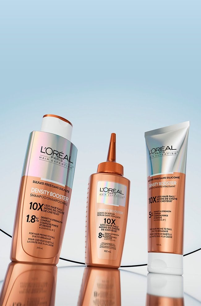 Three L'Oréal Paris Hair Expertise Density Booster products arranged on a blue gradient background with translucent curves. From left to right: a white and copper shampoo bottle, a copper leave-in serum bottle with a thin nozzle, and a copper and white conditioner tube. All products are labeled "Density Booster" and highlight information about reducing hair fall.