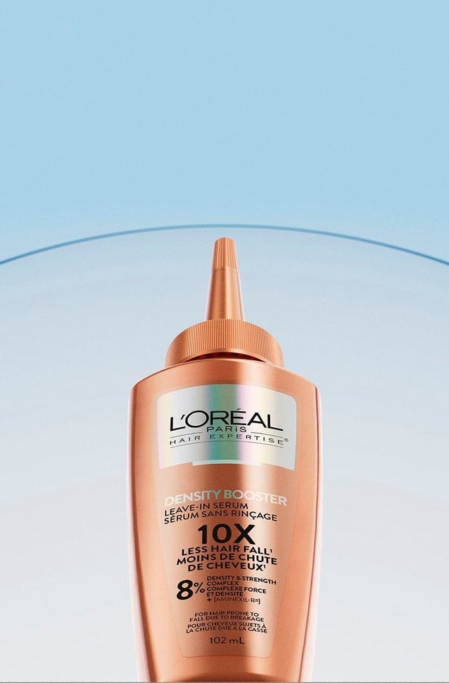 L'Oréal Paris Hair Expertise Density Booster leave-in serum bottle, copper colored with a thin applicator nozzle. The bottle displays "Leave-In Serum", "10x Less Hair Fall", "8% Density & Strength Complex", and "For Hair Prone to Fall" text. It is placed on a blue gradient background with a curved translucent strip