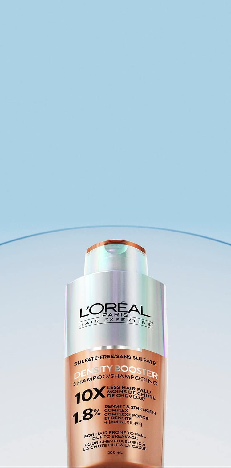 L'Oréal Paris Hair Expertise Density Booster shampoo bottle, copper and white colored, with a golden cap. The bottle is labeled "Sulfate-Free", "10x Less Hair Fall", "1.8% Density & Strength Complex + [Aminexil-R®]", and "For Hair Prone to Fall Due to Breakage". It is positioned on a light blue background with a curved translucent strip in the background.