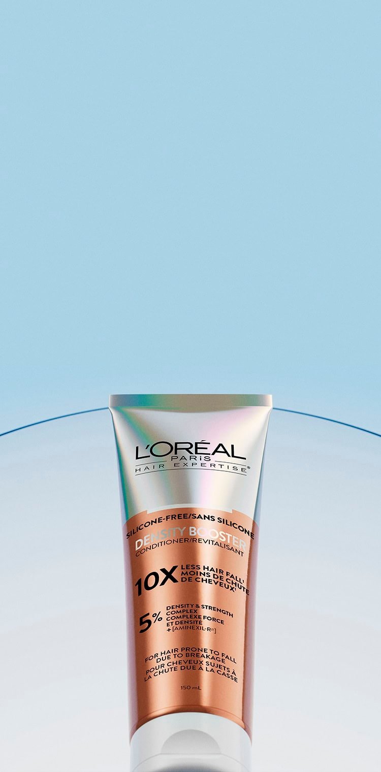L'Oréal Paris Hair Expertise Density Booster conditioner tube, copper and white colored. The tube is labeled "Silicone-Free", "10x Less Hair Fall", "5% Density & Strength Complex", and "For Hair Prone to Fall". It is positioned on a blue gradient background with a curved translucent strip in the background.