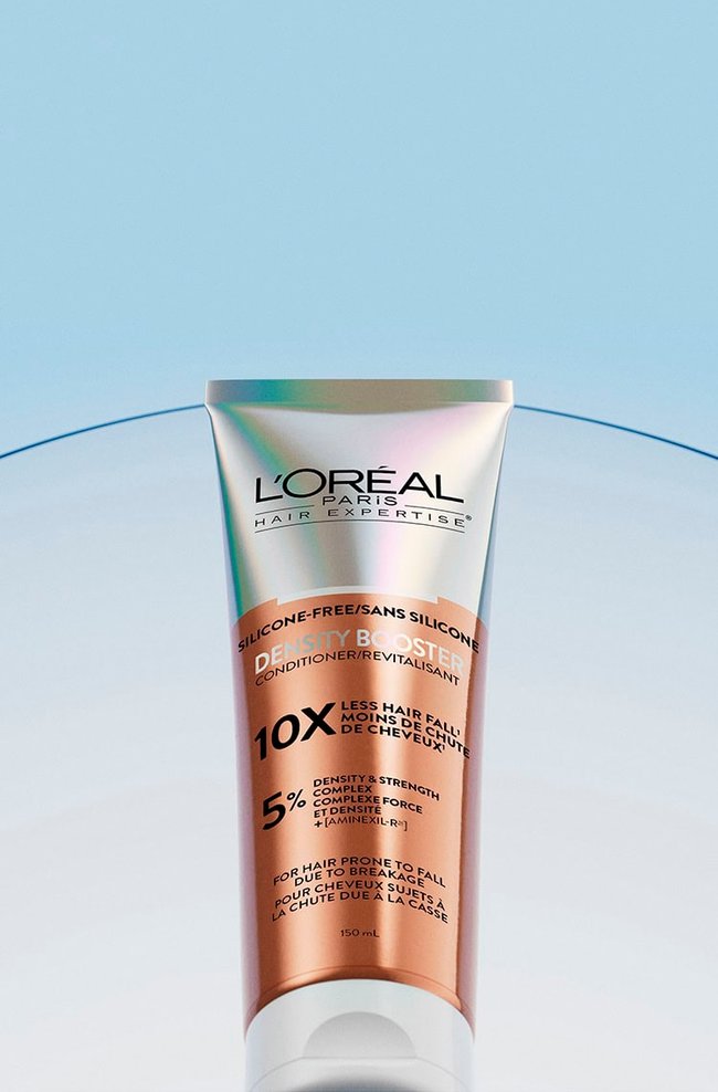 L'Oréal Paris Hair Expertise Density Booster conditioner tube, copper and white colored. The tube is labeled "Silicone-Free", "10x Less Hair Fall", "5% Density & Strength Complex", and "For Hair Prone to Fall". It is positioned on a blue gradient background with a curved translucent strip in the background.