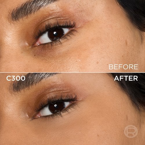 A split image showing a before and after comparison of a person's under-eye and cheek area with medium to dark skin tone. The top section, labeled 'BEFORE', displays skin with slight under-eye discoloration and light eye makeup. The bottom section, labeled 'C300' and 'AFTER', shows the same facial area with noticeably more even-toned, smoother skin, and reduced under-eye circles, following the application of a cosmetic product. The eye makeup remains visible. A subtle 'R' logo is also visible in the bottom right of the 'AFTER' portion.