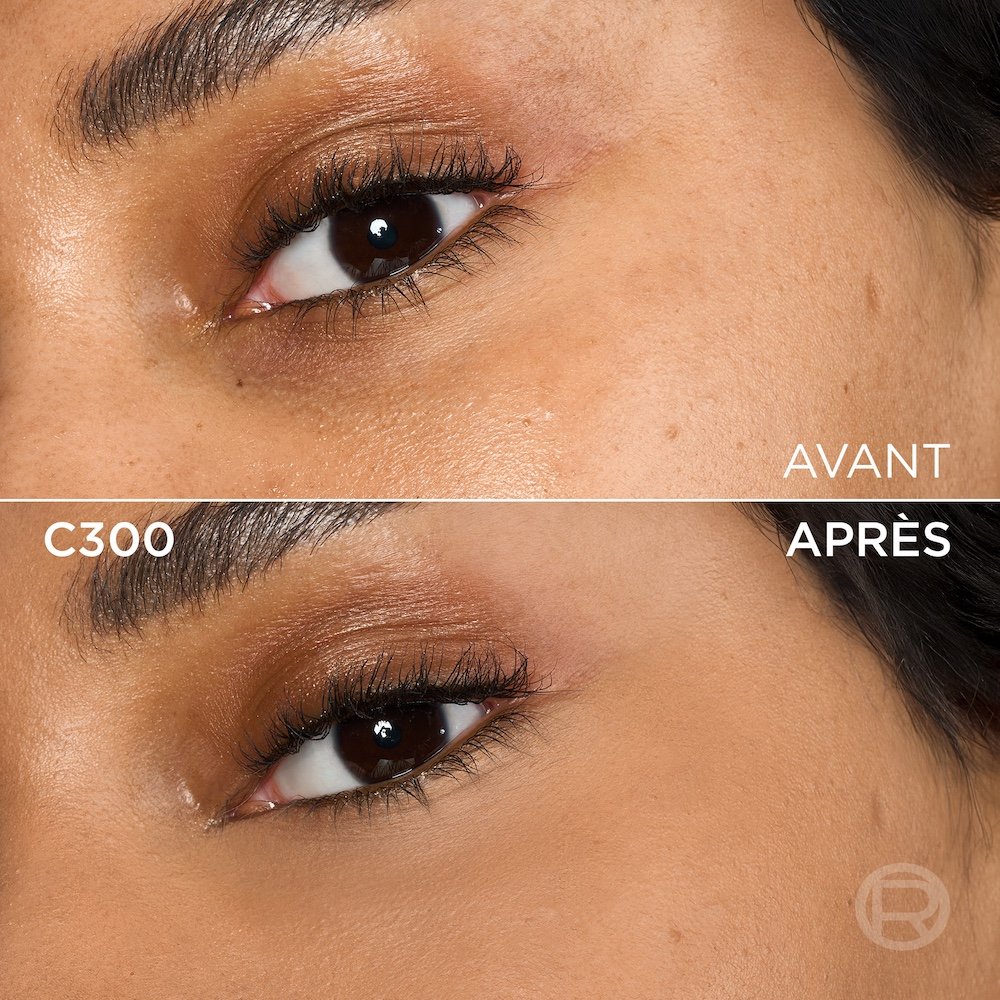 Image divisée montrant un comparatif avant et après de la zone sous l'œil et de la joue d'une personne à la peau de couleur moyenne à foncée. La section supérieure, étiquetée 'BEFORE' (Avant), présente une peau avec une légère décoloration sous l'œil et un maquillage léger des yeux. La section inférieure, étiquetée 'C300' et 'AFTER' (Après), montre la même zone du visage avec une peau visiblement plus unifiée, plus lisse et des cernes atténués, suite à l'application d'un produit cosmétique. Le maquillage des yeux reste visible. Un discret logo 'R' est également visible en bas à droite de la partie 'APRÈS'.