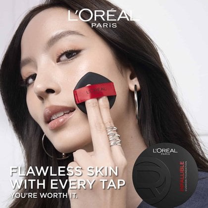 A close-up of a woman with dark hair applying L'Oréal Paris Infallible Cushion Foundation to her left cheek with a black and red cushion applicator. She is wearing a ring with multiple silver bands and a large silver hoop earring. The L'Oréal Paris logo is visible in the top left, and the product packaging for L'Oréal Paris Infallible Cushion Foundation is in the bottom right corner. Overlay text reads 'FLAWLESS SKIN WITH EVERY TAP. YOU'RE WORTH IT