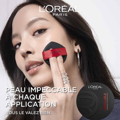 Gros plan sur une femme aux cheveux foncés appliquant le fond de teint L'Oréal Paris Infallible Cushion sur sa joue gauche avec un applicateur coussin noir et rouge. Elle porte une bague à plusieurs anneaux argentés et une grande boucle d'oreille créole argentée. Le logo L'Oréal Paris est visible en haut à gauche, et l'emballage du produit L'Oréal Paris Infallible Cushion Foundation est en bas à droite. Le texte superposé indique 'PEAU IMPECCABLE À CHAQUE APPLICATION. VOUS LE VALEZ BIEN