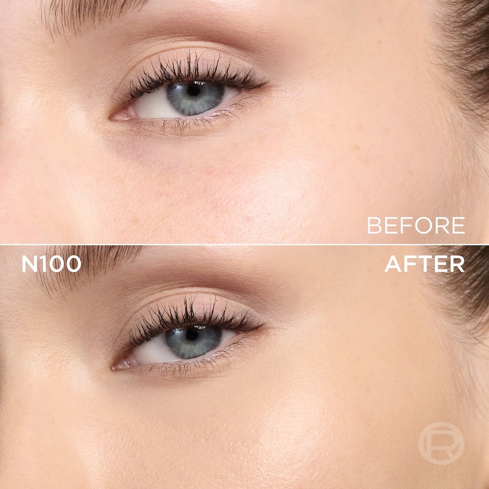A split image showing a before and after comparison of a light-skinned person's under-eye and cheek area with blue eyes. The top section, labeled 'BEFORE', displays skin with a few small spots and slight under-eye discoloration. The bottom section, labeled 'N100' and 'AFTER', shows the same facial area with noticeably more even-toned, smoother skin, and reduced imperfections, following the application of a cosmetic product. A subtle 'R' logo is also visible in the bottom right of the 'AFTER' portion.