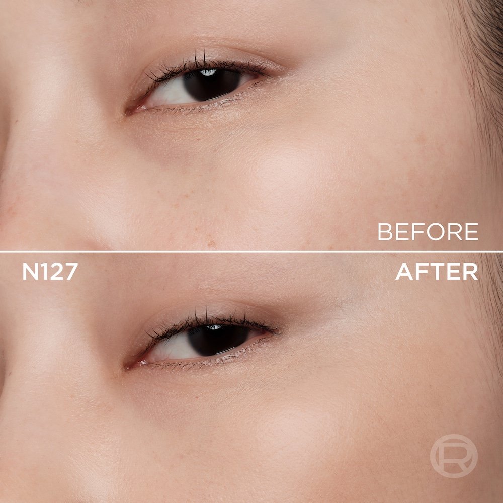A split image showing a before and after comparison of a light-skinned person's under-eye and cheek area. The top section, labeled 'BEFORE', displays skin with a few small freckles and slight under-eye discoloration. The bottom section, labeled 'N127' and 'AFTER', shows the same facial area with noticeably more even-toned, smoother skin, and reduced imperfections, following the application of a cosmetic product. A subtle 'R' logo is also visible in the bottom right of the 'AFTER' portion