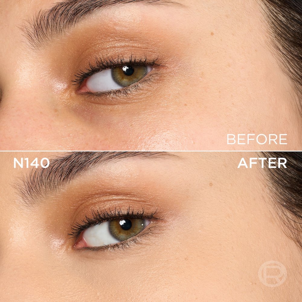 A split image showing a before and after comparison of a light to medium-skinned person's under-eye and cheek area with hazel eyes. The top section, labeled 'BEFORE', displays skin with a few small freckles, visible texture, and light eye makeup including copper eyeshadow. The bottom section, labeled 'N140' and 'AFTER', shows the same facial area with noticeably more even-toned, smoother skin, and reduced imperfections, following the application of a cosmetic product. The eye makeup remains visible. A subtle 'R' logo is also visible in the bottom right of the 'AFTER' portion