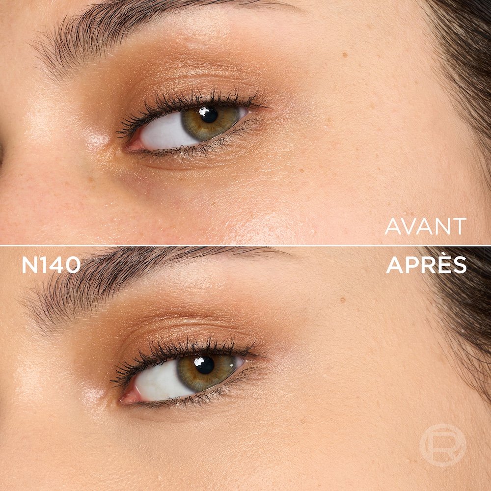 Image divisée montrant un comparatif avant et après de la zone sous l'œil et de la joue d'une personne à la peau claire à moyenne et aux yeux marron-vert. La section supérieure, étiquetée 'BEFORE' (Avant), présente une peau avec quelques petites taches de rousseur, une texture visible et un maquillage léger des yeux avec un fard à paupières cuivré. La section inférieure, étiquetée 'N140' et 'AFTER' (Après), montre la même zone du visage avec une peau visiblement plus unifiée, plus lisse et des imperfections atténuées, suite à l'application d'un produit cosmétique. Le maquillage des yeux reste visible. Un discret logo 'R' est également visible en bas à droite de la partie 'APRÈS'.