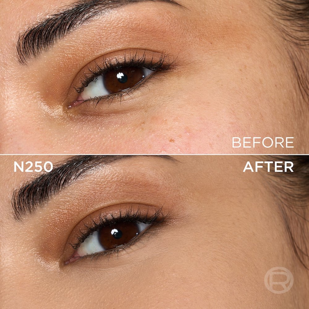 A split image showing a before and after comparison of a medium-skinned person's under-eye and cheek area with brown eyes. The top section, labeled 'BEFORE', displays skin with a few small freckles, visible texture, and light eye makeup including golden eyeshadow. The bottom section, labeled 'N250' and 'AFTER', shows the same facial area with noticeably more even-toned, smoother skin, and reduced imperfections, following the application of a cosmetic product. The eye makeup remains visible. A subtle 'R' logo is also visible in the bottom right of the 'AFTER' portion.