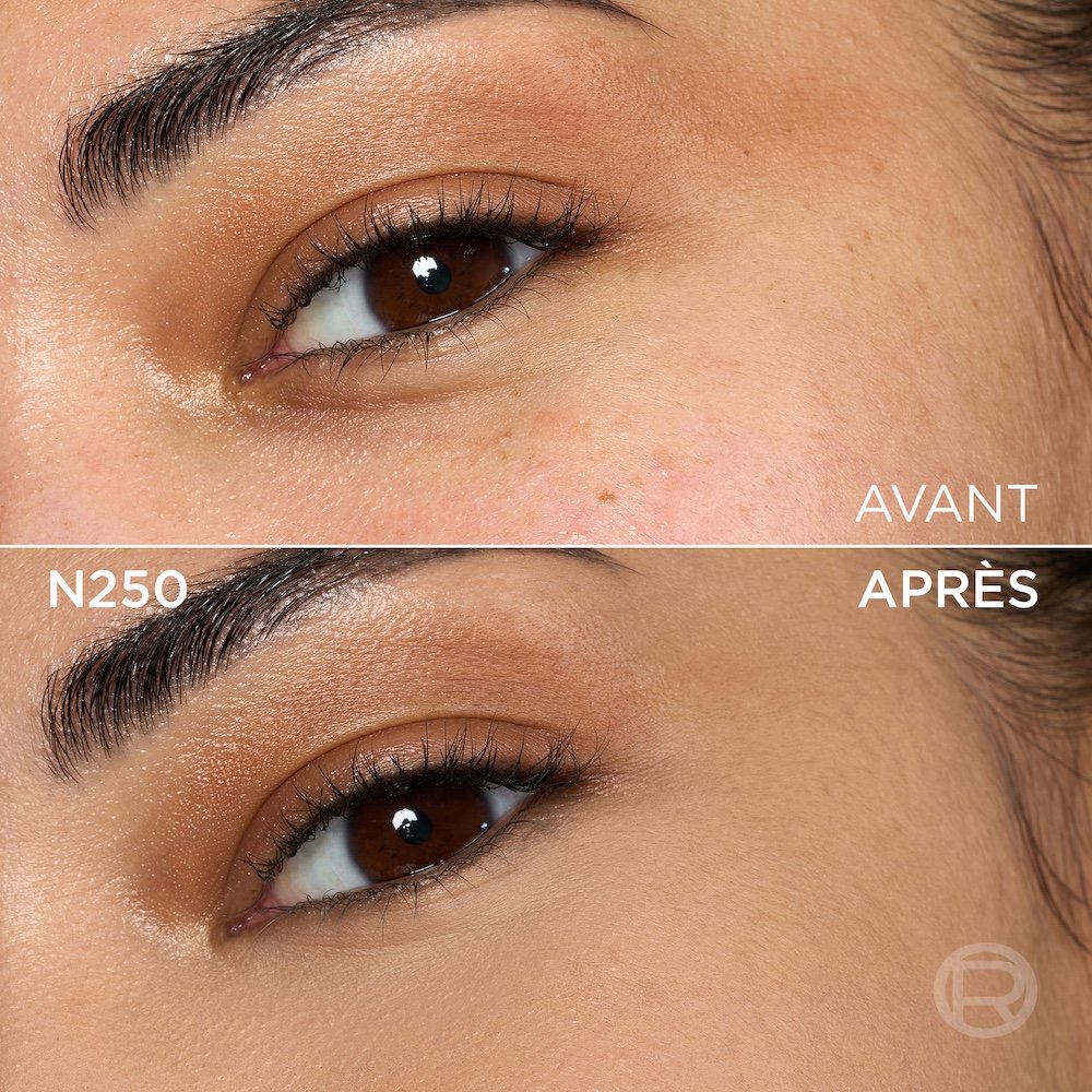 Image divisée montrant un comparatif avant et après de la zone sous l'œil et de la joue d'une personne à la peau de couleur moyenne et aux yeux marron. La section supérieure, étiquetée 'BEFORE' (Avant), présente une peau avec quelques petites taches de rousseur, une texture visible et un maquillage léger des yeux avec un fard à paupières doré. La section inférieure, étiquetée 'N250' et 'AFTER' (Après), montre la même zone du visage avec une peau visiblement plus unifiée, plus lisse et des imperfections atténuées, suite à l'application d'un produit cosmétique. Le maquillage des yeux reste visible. Un discret logo 'R' est également visible en bas à droite de la partie 'APRÈS