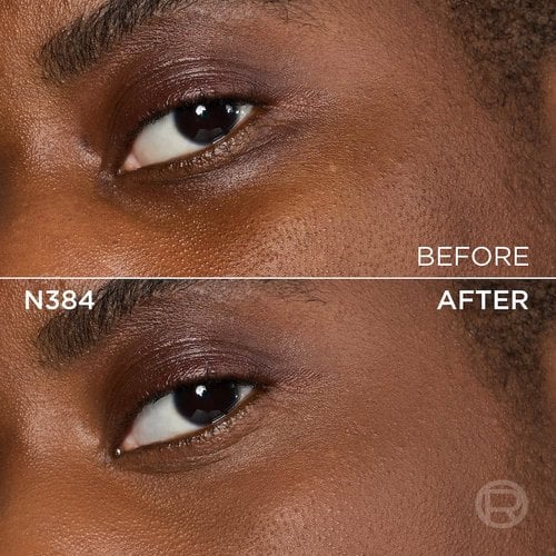 A split image showing a before and after comparison of a very dark-skinned person's under-eye and cheek area with brown eyes. The top section, labeled 'BEFORE', displays skin with visible texture, pores, slight unevenness in tone under the eye, and subtle brown eyeshadow. The bottom section, labeled 'N384' and 'AFTER', shows the same facial area with noticeably smoother, more even-toned skin, and reduced under-eye discoloration, following the application of a cosmetic product. The eye makeup remains similar. A subtle 'R' logo is also visible in the bottom right of the 'AFTER' portion.