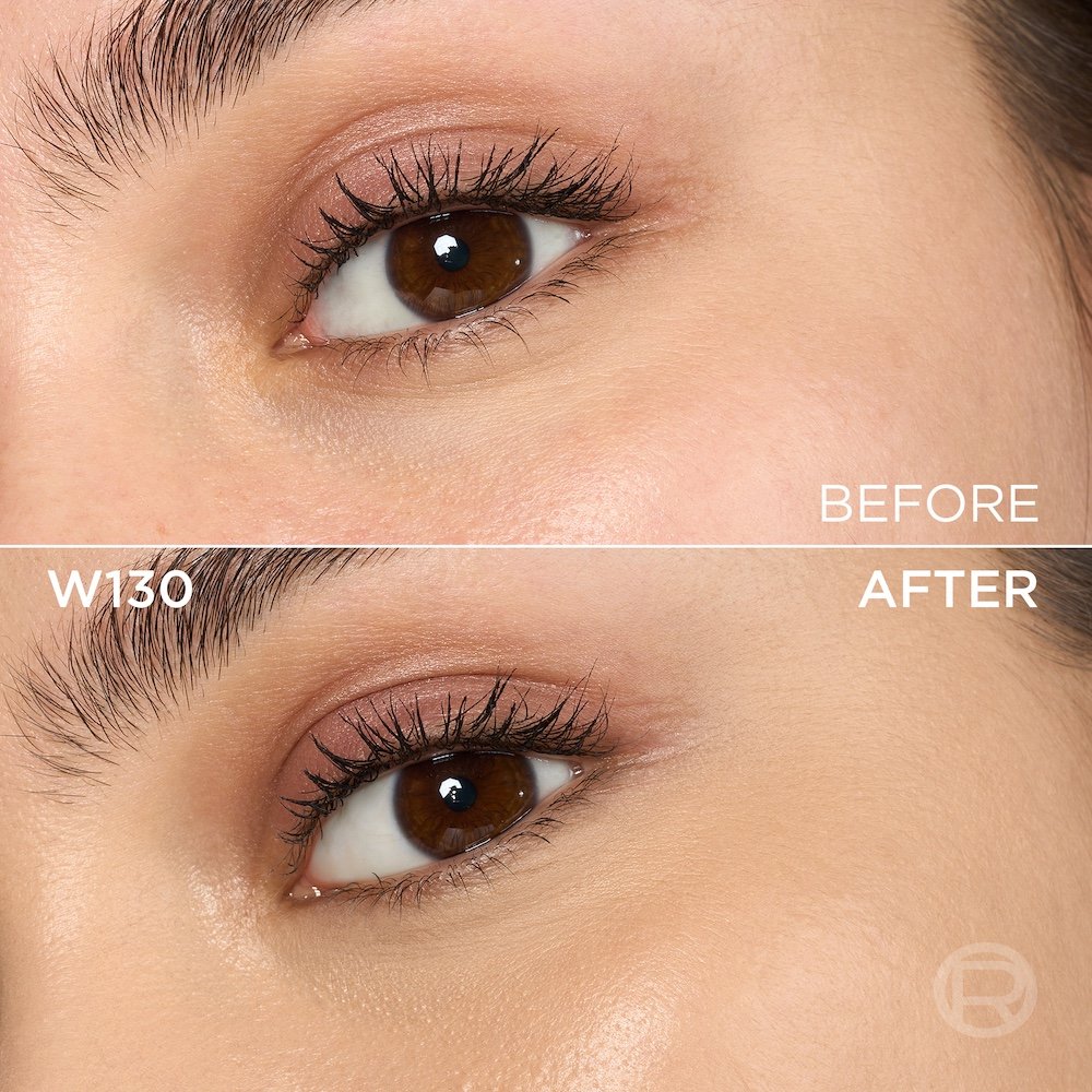 A split image showing a before and after comparison of a light to medium-skinned person's under-eye and cheek area with brown eyes. The top section, labeled 'BEFORE', displays skin with a few slight imperfections and subtle under-eye discoloration, along with light eye makeup featuring pink-brown eyeshadow. The bottom section, labeled 'W130' and 'AFTER', shows the same facial area with noticeably more even-toned, smoother skin, and reduced imperfections, following the application of a cosmetic product. The eye makeup remains visible. A subtle 'R' logo is also visible in the bottom right of the 'AFTER' portion.