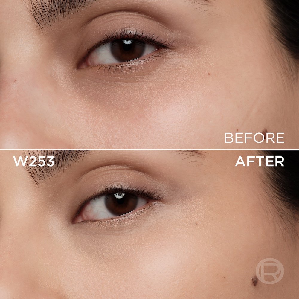 A split image showing a before and after comparison of a light to medium-skinned person's under-eye and cheek area. The top section, labeled 'BEFORE', displays skin with a few slight imperfections, subtle under-eye discoloration, and subtle eye makeup. The bottom section, labeled 'W253' and 'AFTER', shows the same facial area with noticeably more even-toned, smoother skin, and reduced imperfections, following the application of a cosmetic product. The eye makeup remains subtle. A subtle 'R' logo is also visible in the bottom right of the 'AFTER' portion