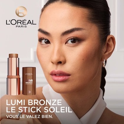Une femme aux cheveux foncés et au teint chaud et olive, regardant directement la caméra, maquillée de manière à rehausser ses traits avec un éclat subtil. Au premier plan, un stick L'Oréal Lumi Bronze et son capuchon sont mis en évidence, avec le texte 'LUMI BRONZE LE STICK SOLEIL VOUS LE VAUTEZ BIEN.' en dessous.