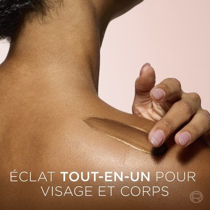 Un gros plan montre l'épaule d'une personne à la peau brune chaude, tandis qu'un produit chatoyant, doré-bronze, est appliqué avec une spatule noire, créant une traînée lisse. Un texte blanc en bas de l'image indique : "ALL-OVER GLOW FOR FACE AND BODY".