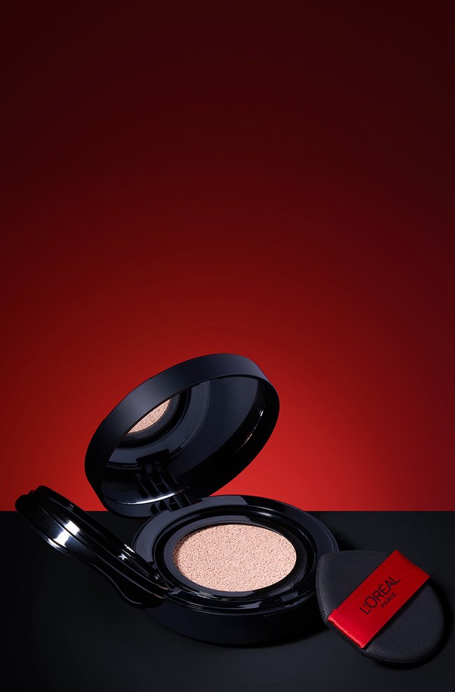 Une photo de produit verticale professionnelle d'un poudrier de fond de teint coussin L'Oréal Paris ouvert avec sa houppette applicatrice sur un fond rouge et noir spectaculaire.