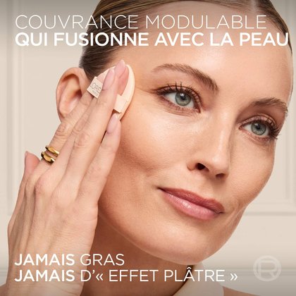 Gros plan d'une femme appliquant le baume teinté sur sa pommette à l'aide de la houppette applicatrice en forme de goutte, avec le texte "BUILDABLE COVERAGE FUSES WITH YOUR SKIN" et "NEVER GREASY NEVER CAKEY".