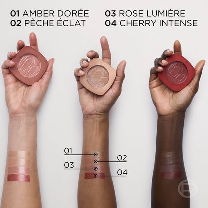 Three hands holding the compacts alongside four color swatches on a forearm, displaying the shade names: "01 AMBER DORÉE", "02 PÊCHE ÉCLAT", "03 ROSE LUMIÈRE", and "04 CHERRY INTENSE".