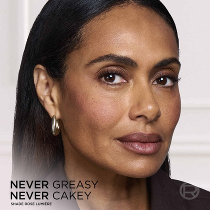 Close-up portrait of a woman showcasing a smooth, flawless, and radiant makeup finish, with the overlay text "NEVER GREASY NEVER CAKEY".