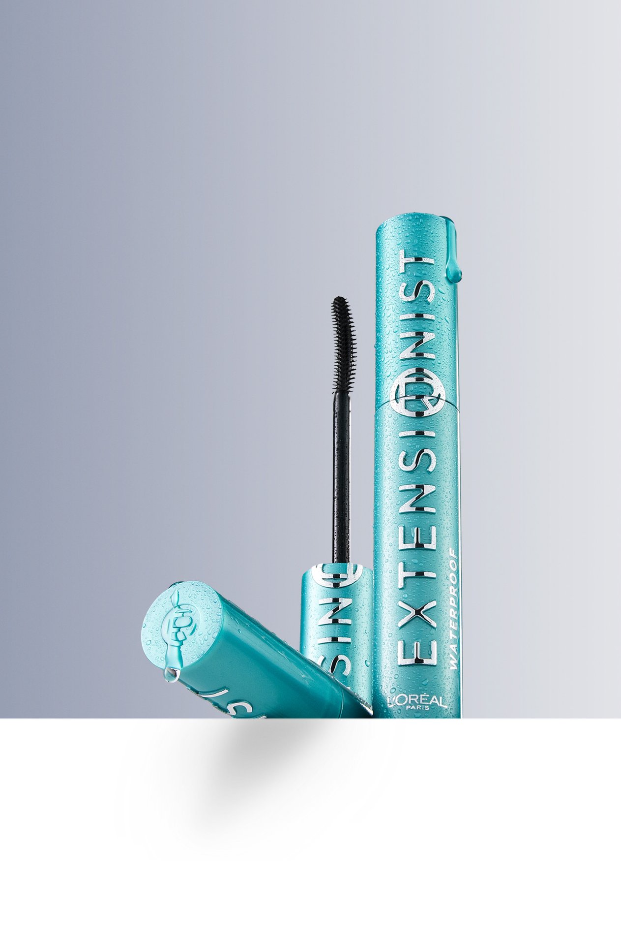 Product photography showing a L'Oréal Paris Telescopic Extensionist mascara in a turquoise tube. The wand stands next to the open tube against a grey-blue gradient background.