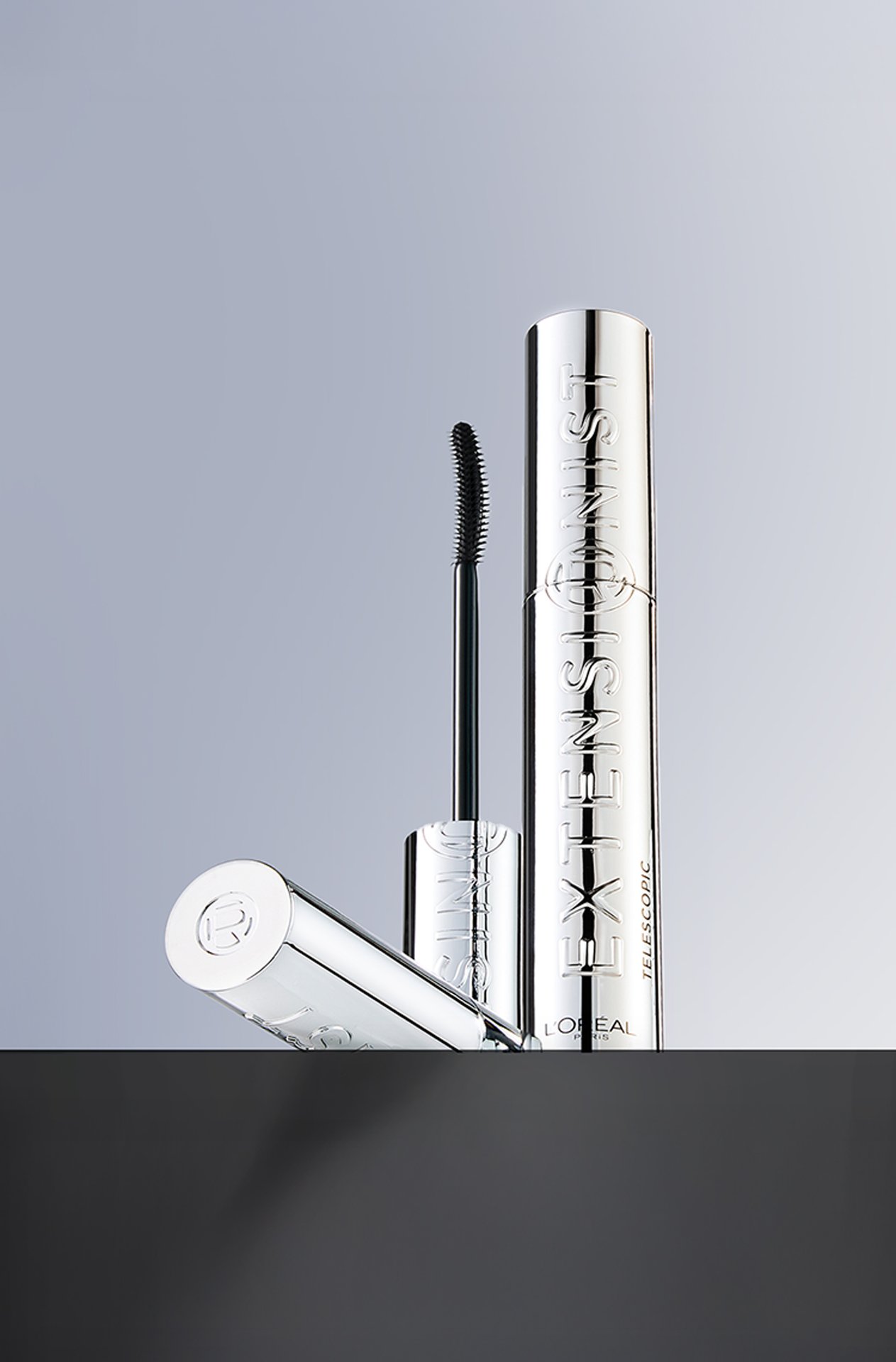 Commercial photography of L'Oréal Paris Telescopic Extensionist mascara with a sleek silver chrome finish. The tube and wand are placed on a shiny black surface in front of a clean grey background.