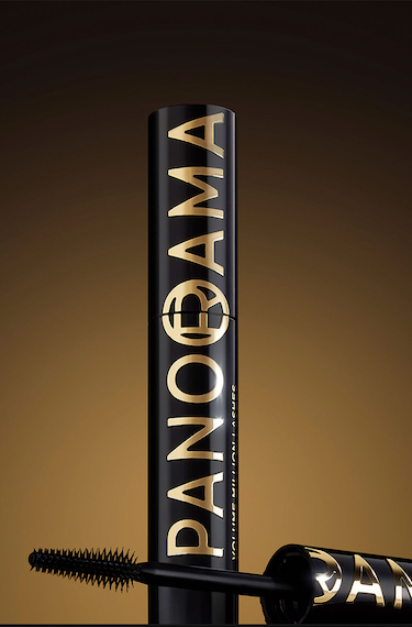 Un tube de mascara noir avec des lettres dorées "PANORAMA" et "VOLUME MILLION LASHES" posé verticalement sur une surface sombre. Le capuchon avec la brosse est allongé à côté du tube, affichant également des lettres dorées. Le fond est un dégradé élégant allant du noir au doré.