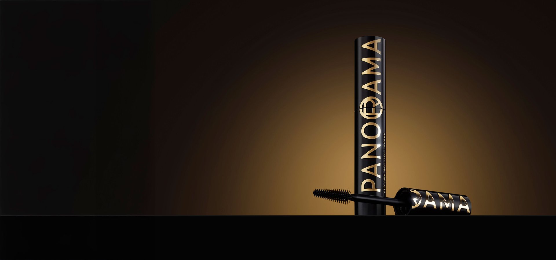 Un tube de mascara noir avec des lettres dorées "PANORAMA" et "VOLUME MILLION LASHES" posé verticalement sur une surface sombre. Le capuchon avec la brosse est allongé à côté du tube, affichant également des lettres dorées. Le fond est un dégradé élégant allant du noir au doré.