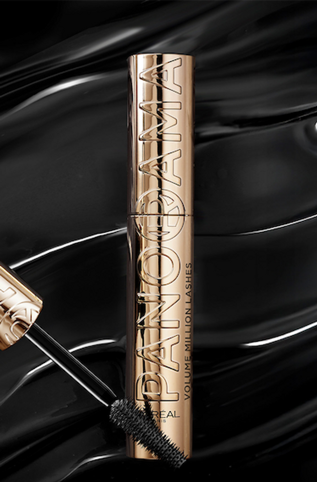 Un tube de mascara doré avec les inscriptions "PANORAMA" et "VOLUME MILLION LASHES" en lettres légèrement en relief, posé verticalement. À côté, le capuchon doré et la brosse noire du mascara sont visibles, avec la brosse s'étendant vers le bas. Le fond est une surface noire brillante et ondulée, qui évoque la texture du mascara ou un effet liquide.