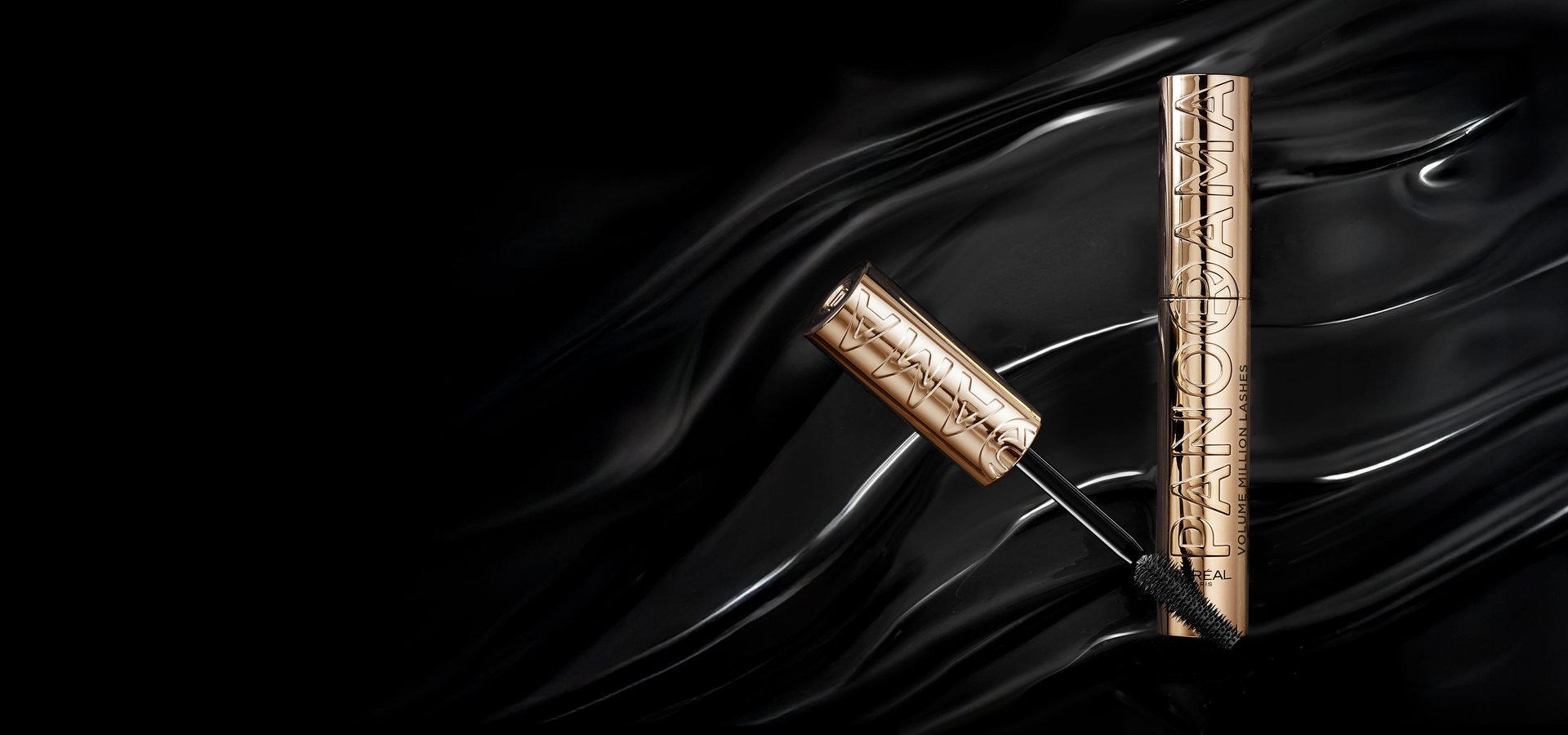 Un tube de mascara doré avec les inscriptions "PANORAMA" et "VOLUME MILLION LASHES" en lettres légèrement en relief, posé verticalement. À côté, le capuchon doré et la brosse noire du mascara sont visibles, avec la brosse s'étendant vers le bas. Le fond est une surface noire brillante et ondulée, qui évoque la texture du mascara ou un effet liquide.​