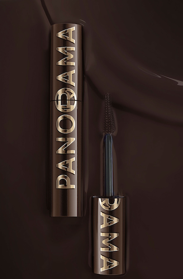 Un tube de mascara de couleur bronze foncé avec l'inscription "PANORAMA" en lettres dorées légèrement en relief, est posé verticalement sur un fond brun chocolat. Le capuchon du mascara est dévissé et positionné en dessous, avec la brosse du mascara visible, également de couleur foncée, s'étendant vers le haut. Le fond présente de légères ondulations qui suggèrent une texture liquide ou veloutée.