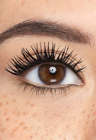 A close-up shot of a person's open eyes featuring dramatically volumized and lengthened eyelashes, coated with mascara. The person has light brown eyes, well-groomed eyebrows, and visible freckles across their nose and cheeks, showcasing the effect of the mascara.
