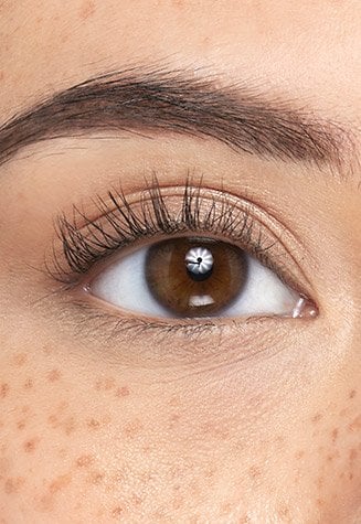 A close-up shot of a person's open eyes with natural, subtle eyelashes. The person has light brown eyes, neatly groomed eyebrows, and visible freckles across the bridge of their nose and under their eyes.