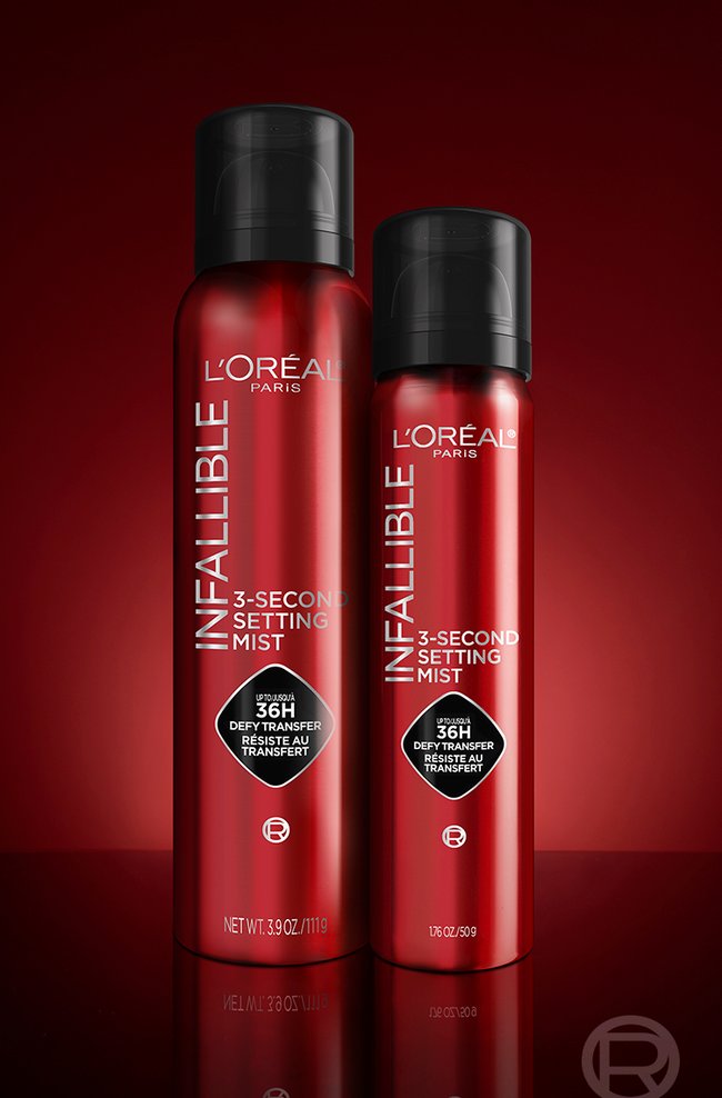 Deux flacons vaporisateurs rouges de "L'Oréal Infallible 3-Second Setting Mist" sont présentés sur une surface noire brillante avec un fond dégradé rouge. Le flacon de gauche est plus grand (3,9 oz / 111 g), tandis que celui de droite est plus petit (1,7 oz / 50 g). Les deux flacons sont dotés de capuchons noirs et d'un branding qui met en évidence "Up to 36H transfer-proof / Defy transfer / Résiste au transfert".
