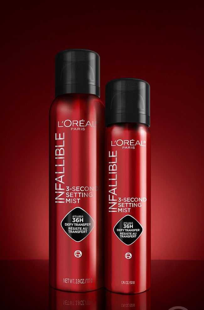 Two red spray bottles of "L'Oréal Infallible 3-Second Setting Mist" are displayed on a glossy black surface with a red gradient background. The bottle on the left is larger (3.9 oz / 111 g), while the one on the right is smaller (1.7 oz / 50 g). Both bottles feature black caps and branding that highlights "Up to 36H transfer-proof / Defy transfer / Résiste au transfert."