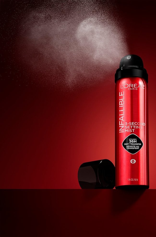 The red L'Oréal Infallible 3-Second Setting Mist spray bottle, with its cap off, releasing a fine mist against a deep red background.