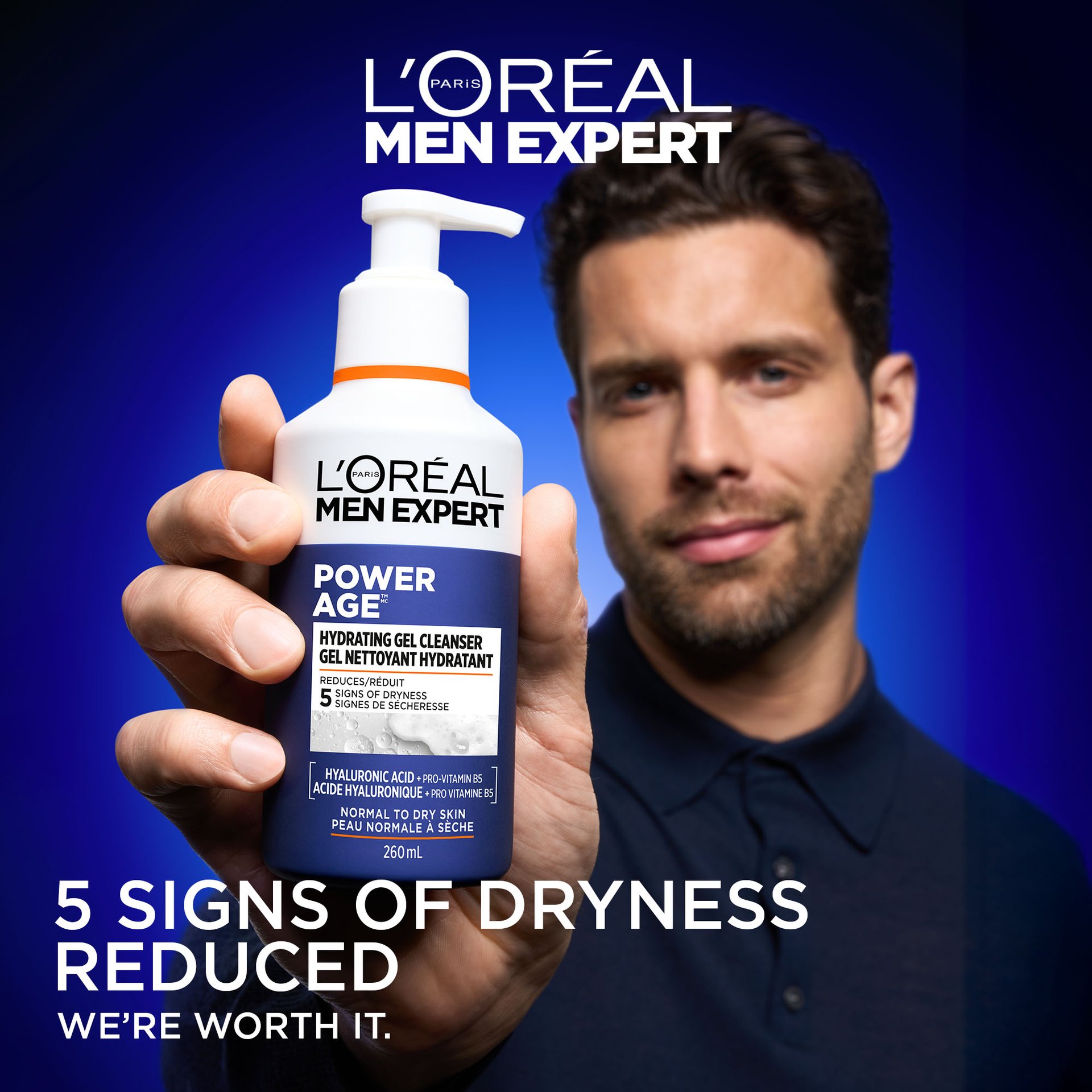 A man with short dark hair and a beard holds the Power Age Hydrating Gel Cleanser bottle close to the camera. The background is a vibrant gradient blue. Large text at the bottom reads "5 SIGNS OF DRYNESS REDUCED" with the L'Oréal slogan "WE’RE WORTH IT."