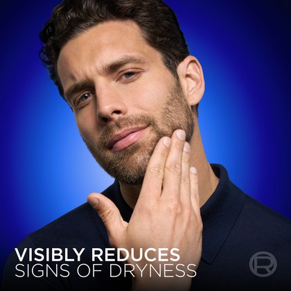 A close-up shot of the same bearded model from Image 2, touching his cheek with his hand to show smooth skin. The background is dark blue. Text at the bottom states: "VISIBLY REDUCES SIGNS OF DRYNESS."