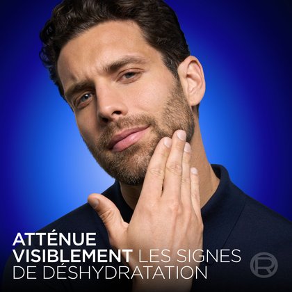 Un gros plan du même mannequin barbu de l'image 2, touchant sa joue avec sa main pour montrer une peau lisse. Le fond est bleu foncé. Le texte en bas indique : « RÉDUIT VISIBLEMENT LES SIGNES DE SÉCHERESSE ».