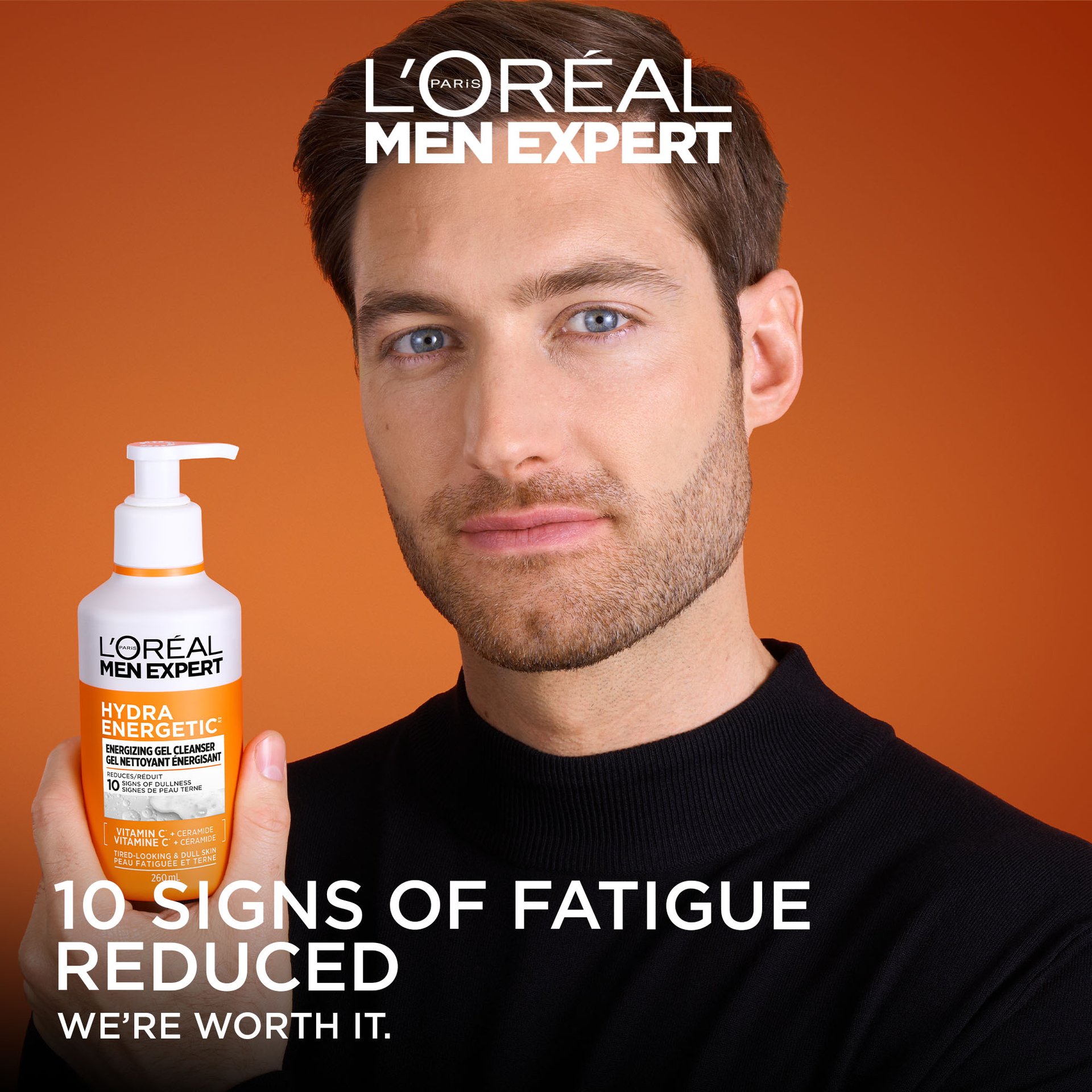  A man with groomed facial hair holds the Hydra Energetic Energizing Gel Cleanser bottle. The background is a warm gradient orange. Text at the bottom reads "10 SIGNS OF FATIGUE REDUCED" with the L'Oréal slogan "WE’RE WORTH IT."
