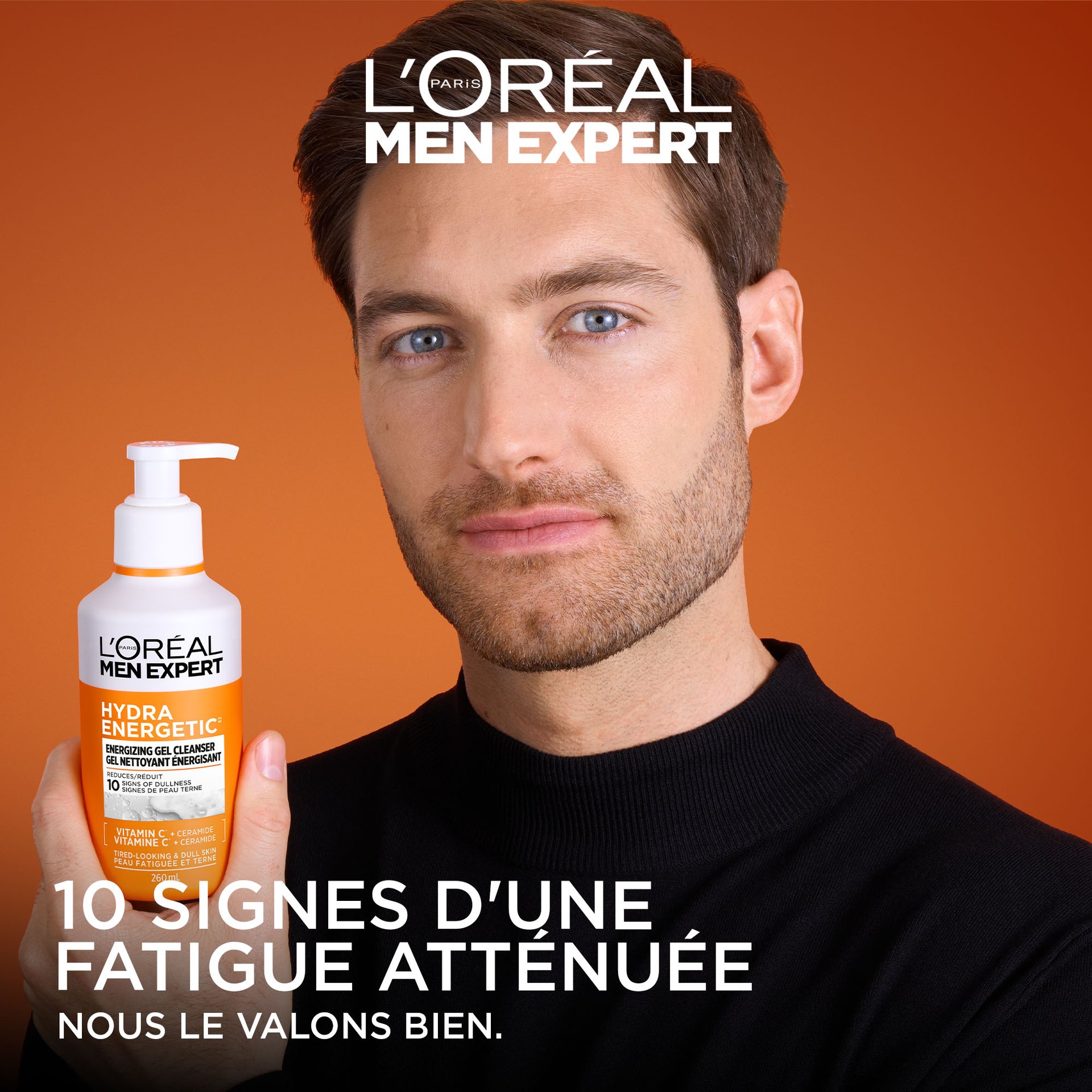 Un homme avec une barbe soignée tient le flacon de Gel Nettoyant Énergisant Hydra Energetic. Le fond est un dégradé d'orange chaud. Le texte en bas indique « 10 SIGNES DE FATIGUE RÉDUITS » avec le slogan de L'Oréal « PARCE QUE NOUS LE VALONS BIEN ».