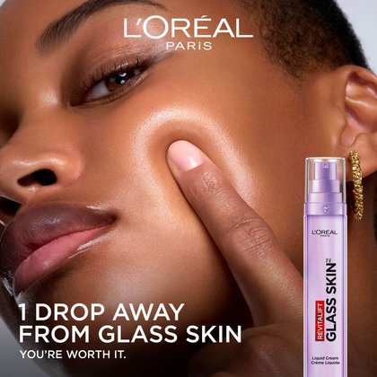 A close-up of a woman touching her radiant, dewy cheek to show the product's luminous effect. The purple pump bottle is pictured next to her with the English text: "1 Drop Away From Glass Skin."